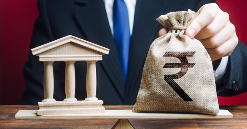 Mérlegen álló oszlopos épületmodell és pénzeszsák, üzleti környezetben.