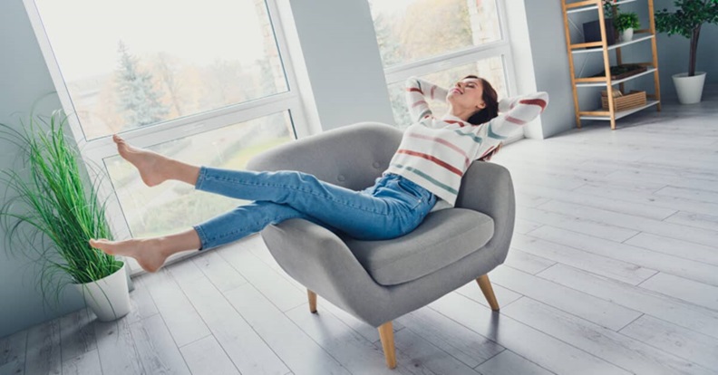 Egy személy kényelmesen hátradől egy világos, modern nappaliban elhelyezett fotelben, lábait a karfára téve, nyugodt és pihentető környezetben.