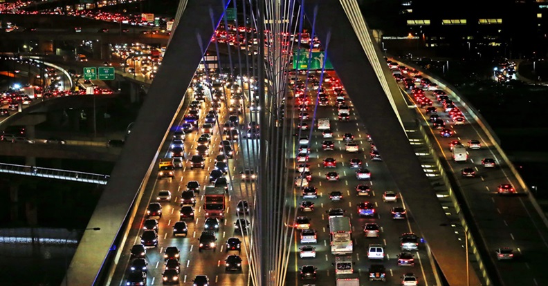 Éjszakai felülnézeti kép egy forgalmas autópálya hídról, sűrűn egymás mellett haladó autósorokkal és városi fényekkel, a zsúfolt közlekedést szimbolizálva.