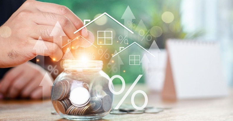 Kéz egy pénzzel teli befőttesüveg fölött, háttérben házakat és százalékjeleket ábrázoló grafikus ikonokkal, a megtakarítás és befektetés növekedésének szimbolikus megjelenítéseként.