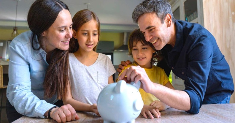 Egy család közösen tesz pénzt egy fehér perselymalacba az asztalnál, miközben a szülők a gyerekeknek mutatják a megtakarítás folyamatát.